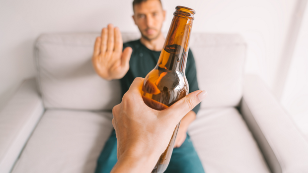 Ein junger Mann sitzt auf einem hellen Sofa und lehnt mit ausgestreckter Hand eine Flasche Bier ab, die ihm gereicht wird.